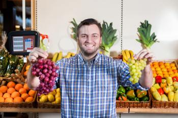 Portrait of smiling man in shirt selling ripe grapes in shop. Boxes with fruits and vegetables on the background. 