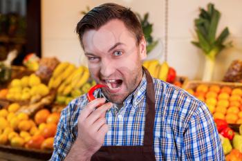 Portrait of man in shirt and apron trying to eat a chili pepper in supermarket. Boxes with fruits and vegetables on the background. 
