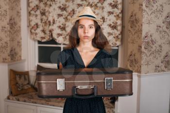 Young beautiful woman holding suitcase in her hands. Vintage style. Sofa and window with curtains on the background.