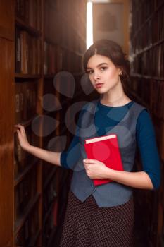 Young girl with the book in hands standing between book shelves