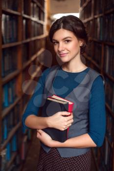 Smiling student girl or woman with books between bookshelves in library. 