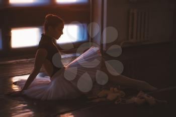 Ballerina sitting on the floor in dark ballet class against window full of sunlight.