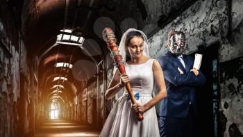 Newlyweds maniacs, an interior of old prison on background. Couple with bloody bat and meat cleaver, groom in hockey mask