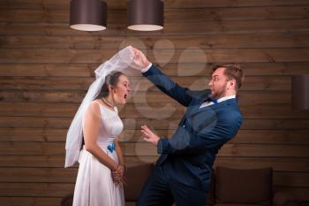 Young bride in white dress and veil against serious groom in suit, wooden background