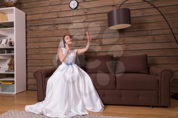 Portrait of bride in white dress talking by mobile phone and looking at wedding ring on her hand, wooden room interior on background.