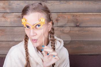 Young woman with yellow glasses on a stick, wooden background. Funny photo props. Fun accessories for shoots