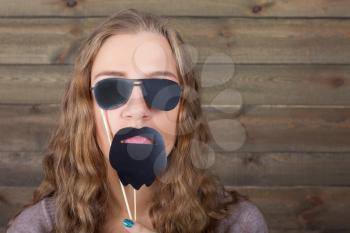 Playful woman with funny sunglasses and beard on a stick, wooden background. Fun photo props and accessories for shoots