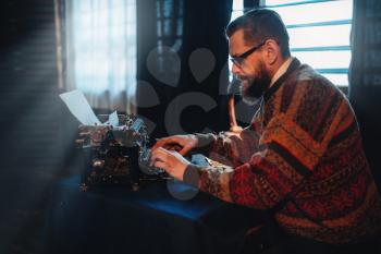 Bearded writer in glasses typing on retro typewriter against window with sunlight