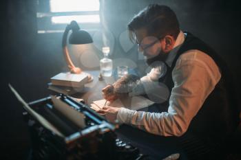Bearded writer in glasses writes novel with a feather in a smoky room.  Retro typewriter, crystal decanter, books and vintage lamp on the desk