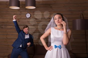 Portrait of bride with mobile phone against happy groom, wooden room on background.