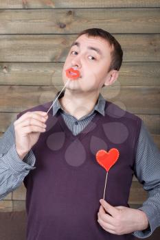 Young man with funny red lips and heart on a sticks in hand, wooden background. Fun photo props and accessories for shoots