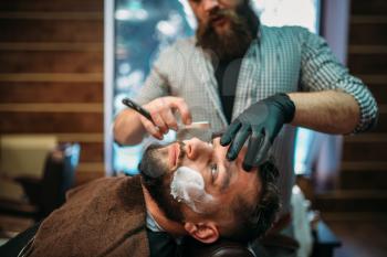 Barber shaves the beard of the client by straight razor at barbershop.