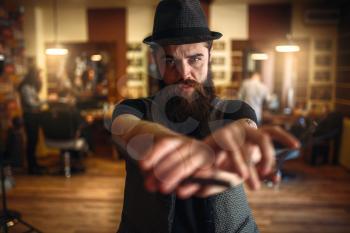 Brutal bearded barber in vintage hat holding scissors and straight razor in hands, selective focus, blur background
