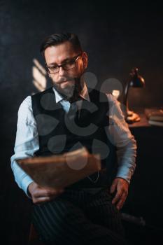 Portrait of seriuos bearded man in glasses reads handwritten text. Desk with retro lighting lamp on background. Writer, journalist, literature author, blogger or poet concept