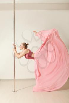Slim professional poledance girl posing in beautiful pink dress. Sexy strip dancer exercising with pole in dance studio