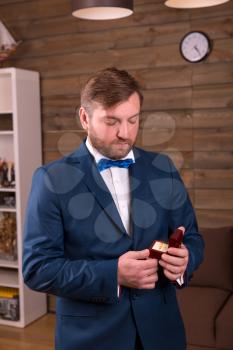 Success groom in luxury suit holding box with wedding ring in his hand, wooden room interior on background