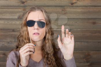 Playful girl with funny sunglasses on a stick represents the blind person, wooden background. Fun photo props and accessories for shoots