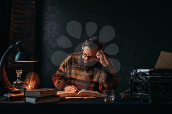Bearded author in glasses reading a book. Retro typewriter, feather, crystal decanter, books and vintage lamp on the desk