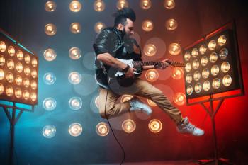 Male performer with electro guitar in a jump on the stage with the decorations of lights. Music entertainment. Bearded musican song performing 