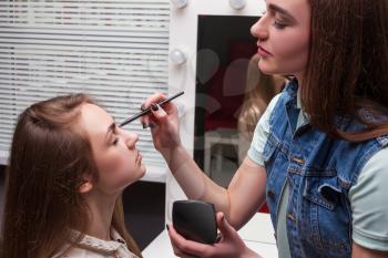 Female make up artist with brush in hands applying make-up on beautiful girl face, beauty studio on background. Cosmetic salon