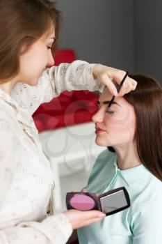 Female make up artist with brush in hands applying make-up on beautiful girl face, beauty studio on background. Cosmetic salon