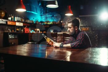 Sad man sitting at the bar counter and holds the bottle with alcohol beverage in hands. Male person in pub, alcoholism, alcohol addiction, drunkenness