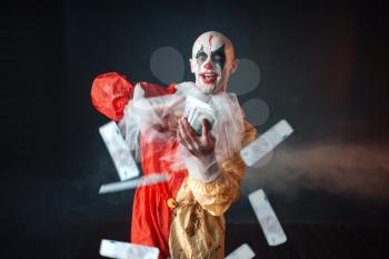 Scary bloody clown with crazy eyes holds fan of money. Man with makeup in carnival costume, mad maniac