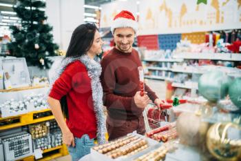 Young couple with cart in department of holiday decorations in supermarket, family tradition. December shopping of new year or christmas goods