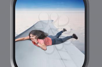 Communication addiction, young woman with phone lies on plane wing  in flight, sky and clouds on background