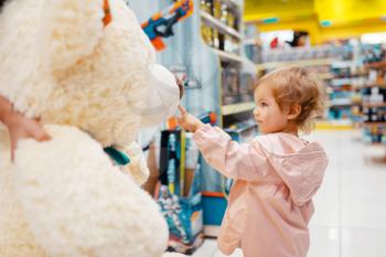Little girl choosing big teddy bear in kids store, side view. Daughter looking for toys in supermarket, family shopping, young customer