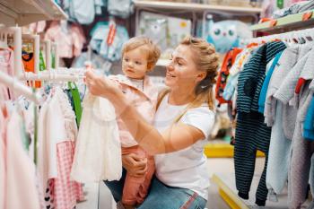 Mother with her little girl choosing clothes in kids store. Mom and child buying dress in supermarket together, family shopping