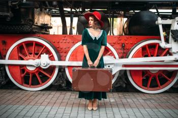 Woman in red hat with suitcase in hands against vintage steam locomotive, red wheels closeup. Old train. Railway engine, railroad journey