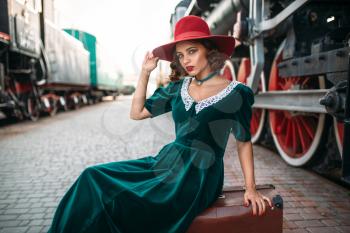 Woman in red hat sitting on suitcase against vintage steam train. Old locomotive. Railway engine, ancient railroad vehicle