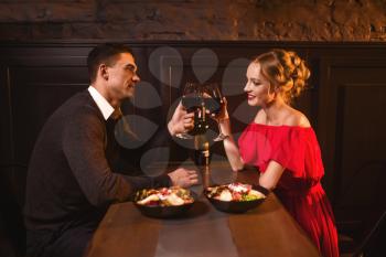 Beautiful love couple raised glasses with red wine in restaurant, romantic date. Elegant woman in red dress and her man, anniversary celebration