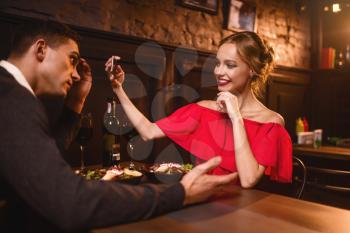 Smiling woman in red dress makes image of her man on phone camera. Beautiful love couple in restaurant, romantic evening, anniversary celebration