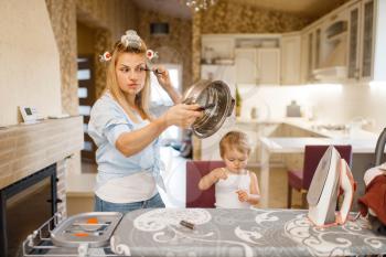 Housewife paints eyelashes at the ironing board, little kid looks at her. Woman with child at home together