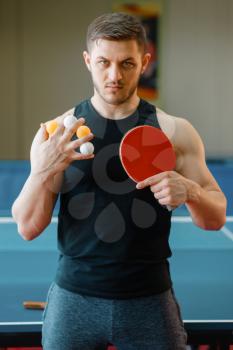 Man with racket holds table tennis balls between fingers, workout indoors. Male person in sportswear standing at the table with net, training in ping pong club