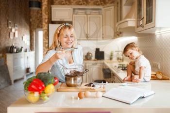 Young mother and her daughter tastes melted chocolate. Cute woman and little girl cooking on the kitchen, pastry preparation. Happy family prepares sweet dessert at the counter