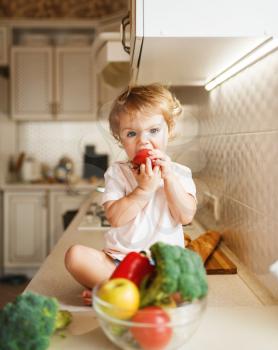 Little girl sitting on the table and eats tomato. Female baby tasting vegetables from the bowl on the kitchen. Child tastes vegetables