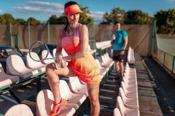 Attractive woman with tennis racket poses on the podium, male partner on background. Summer season sport game. Active lifestyle