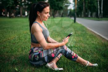 Slim woman in headphones sitting on grass and relax. Sporty girl on outdoors fitness workout