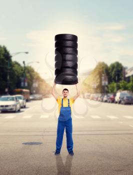 Tyre service worker in uniform holds pile of tires over head. Repairman, wheel mounting