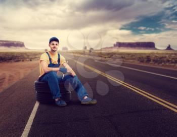Service mechanic in uniform sitting on tire, desert road on background. Repairman, wheel mounting, tyre service