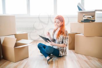Young woman with notebook sitting on the floor among cardboard boxes, housewarming. Relocation to new home