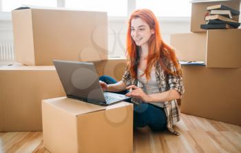 Smiling girl using laptop among cardboard boxes, moving to new house, housewarming
