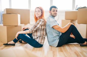 Happy couple sitting on the floor with their backs to each other among cardboard boxes, moving to new home, housewarming