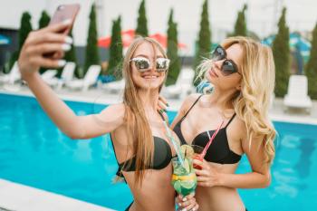 Two sexy girls in swimsuits and sunglasses makes selfie near the swimmimg pool. Slim women sitting by the poolside, resort holidays
