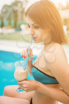 Sexy woman sitting by the poolside and drinks cocktail. Happy girl relax near the swimming pool. Summer vacations
