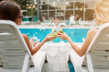 Two sexy girls relax with cocktails on deck chairs near the swimmimg pool. Slim women sitting by the poolside, resort holidays 