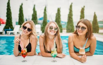 Three happy girls in swimsuits and sunglasses drinks cocktails on the poolside. Resort holidays. Tanned women relax in the swimmimg pool
